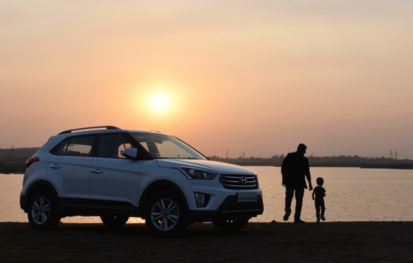 Vehicle parked by pond. Parent and child are standing next to it watching the sunset.