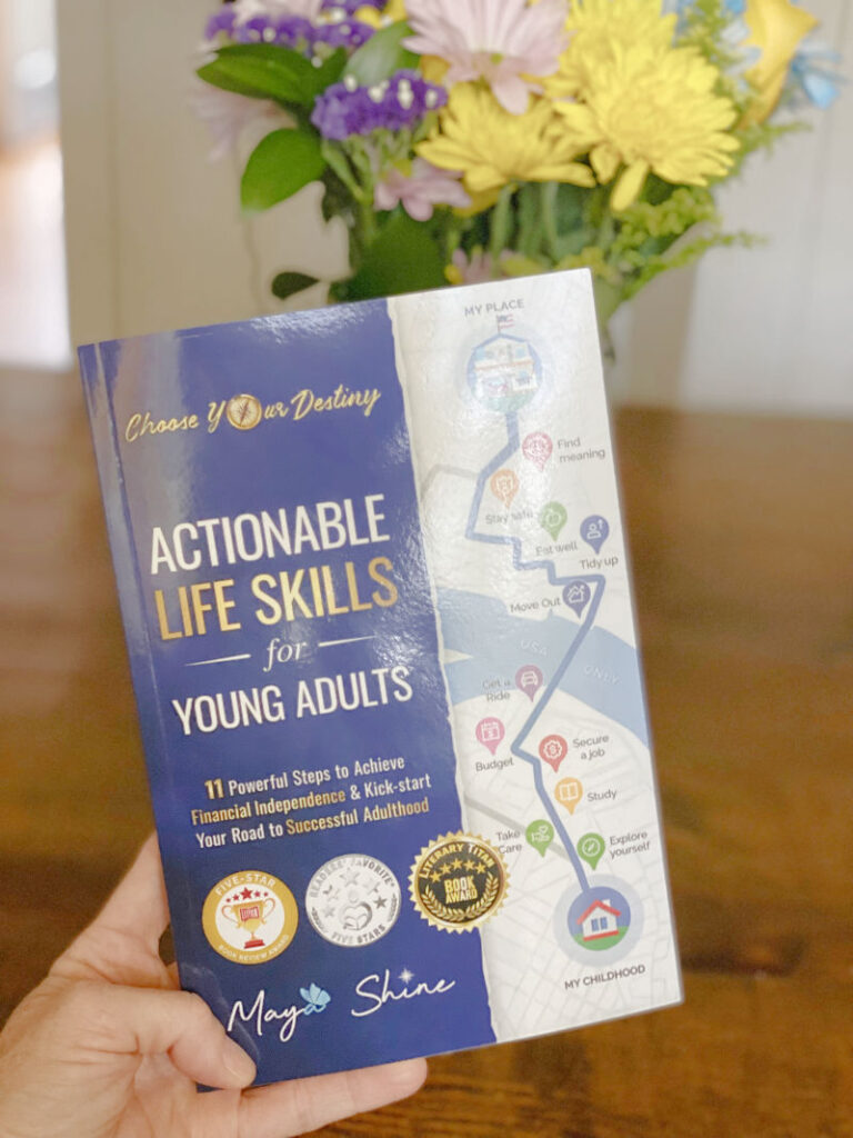 Book being held up with vase of flowers behind it. Title of book reads Actionable Life Skills for Young Adults by Maya Shine.