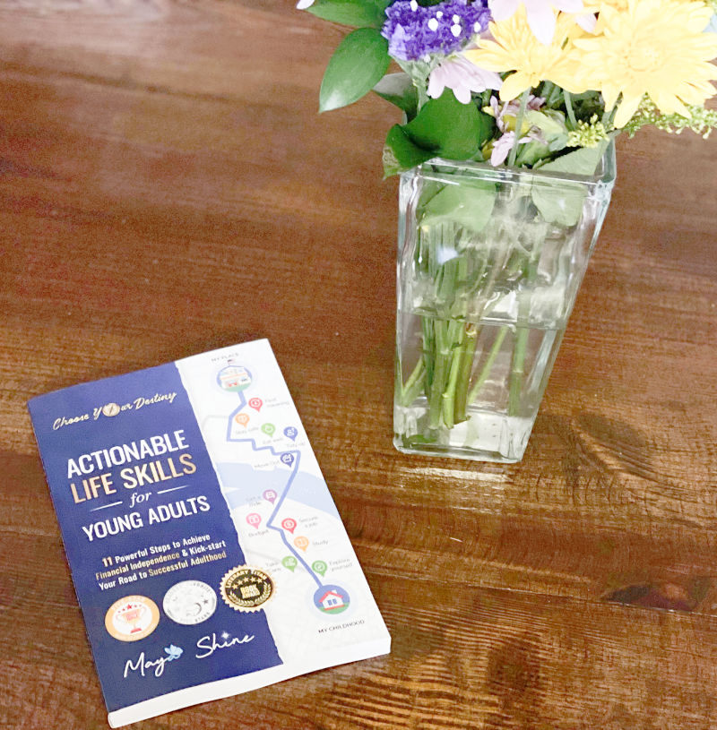 Book entitled Actionable Life Skills for Young Adults is on the kitchen counter with a vase of flowers next to it.