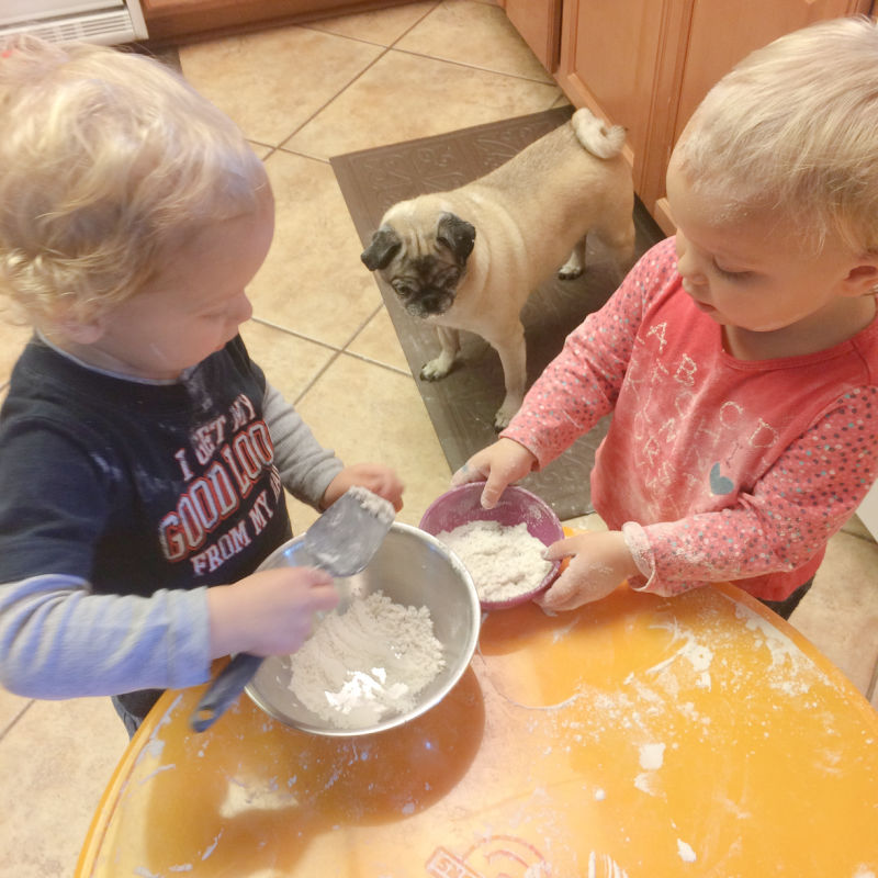 Twin toddlers standing around a child's play table handling flour in a bowl and with kitchen utensils for a sensory bin for toddlers.