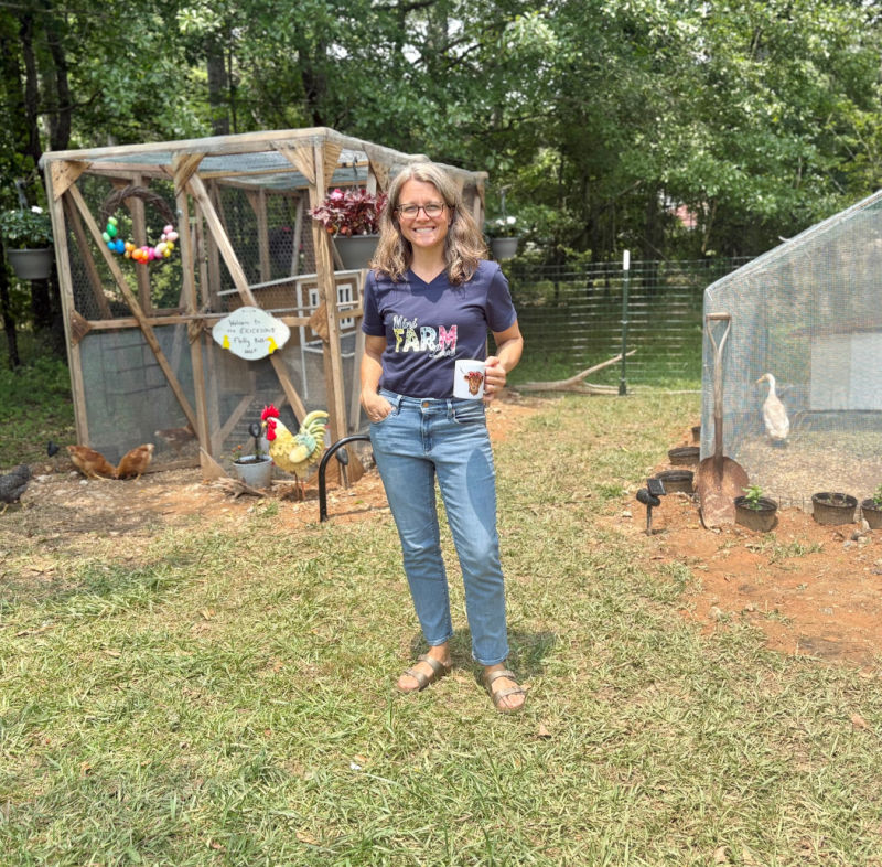 Jaimi Erickson standing in front of her mini farm wearing mini farm love t-shirt and holding Petunia mug from Katie VanSlyke's farm swag collection.